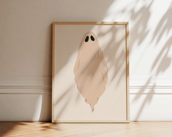 May include: A framed wall art print featuring a beige ghost illustration with two black oval eyes centred on a light beige background. The artwork is enclosed in a simple light-coloured wooden frame, presenting a clean and neutral aesthetic.