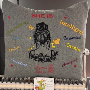 May include: Grey cushion with embroidered text and a woman's silhouette. The cushion features the words "SHE IS" in red, and other words like "Magical", "Intelligent", and "Amazing" in various colours. The phrase "I am She, She is Me" is also embroidered.