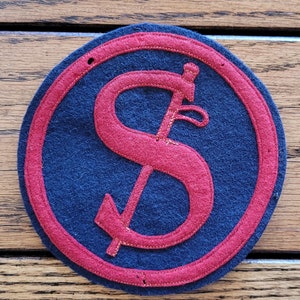 May include: A red felt patch with a stylized red dollar sign on a dark blue background. The patch has a red border.