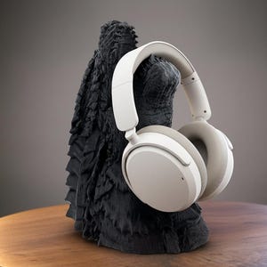 Puede incluir: Auriculares blancos y beige sobre la oreja colocados en un soporte negro texturizado. El soporte tiene una apariencia rugosa y tallada. Los auriculares son de diseño moderno.