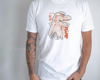May include: White t-shirt featuring a Godzilla graphic in orange and black. The design includes Japanese text and the years 1954-1975. The graphic depicts Godzilla's head and a cityscape.