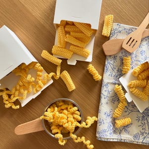 May include: A top-down view displays handmade crochet food items. Yellow crochet pasta, including rigatoni and fusilli, spills from white boxes and a small metal bowl with a wooden handle. Wooden spoons and a floral patterned cloth add to the kitchen theme.