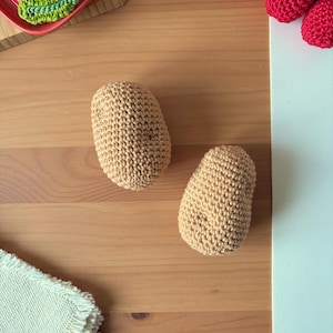 Peut inclure: Deux objets beiges en forme de pomme de terre crochetés sont présentés sur une surface en bois. L'image montre également des fruits crochetés, dont un citron, une orange et des cerises, suggérant un projet artisanal sur le thème de l'alimentation.