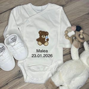 Könnte beinhalten: Weißer Baby-Body mit einem braunen Teddybär-Applikation, der eine Flasche hält. Auf dem Body stehen der Name "Malea" und das Datum "23.01.2026". Inklusive weißer Babyschuhe, einem Beißring aus Holz und einem Stofflamm.