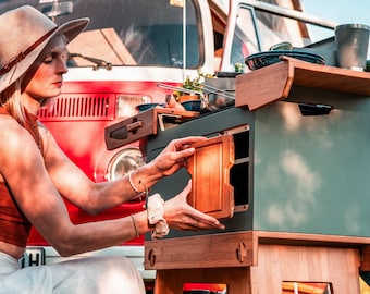 May include: A woman opens a wooden drawer on a portable outdoor kitchen. The kitchen has a green painted surface, a wooden countertop, and a shelf with a metal cup and a lantern.