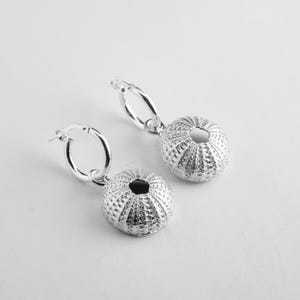 Pode incluir: Um par de brincos de argola em prata com pingentes em forma de conchas de ouriço-do-mar. Cada pingente tem uma superfície esférica texturizada com uma pequena abertura circular no topo, suspenso por uma argola de prata polida simples.
