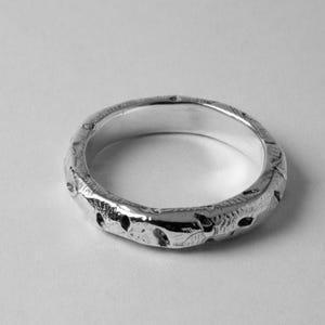May include: A silver ring with a textured, patterned design. The ring is smooth on the inside and has a slightly rounded shape.