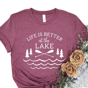May include: A maroon t-shirt with the text "Life is better at the lake" and a graphic of two trees, two paddles crossed, and wavy lines. The shirt is tied in a knot at the bottom.