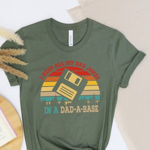May include: A green t-shirt with a retro sunset graphic and a floppy disk. The text on the shirt reads "I keep all my dad jokes in a dad-a-base".