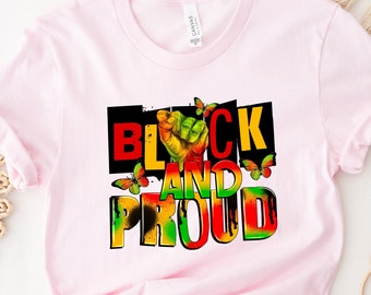 May include: A light pink t-shirt with the words "BLACK AND PROUD" in a colorful graphic design. The design includes a raised fist and butterflies. The shirt is made of a soft material and has short sleeves.