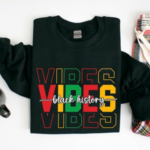 May include: Black sweatshirt with the text "VIBES black history VIBES" in red, green, and yellow lettering. The sweatshirt is folded and laid flat on a white surface.
