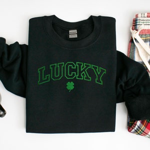 以下が含まれることがあります： 緑のクローバーと、その上に緑色のアーチ状に「LUCKY」と書かれた黒いスウェットシャツ