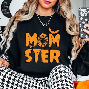 K&ouml;nnte beinhalten: Schwarzer Pullover mit orangefarbenem Text, der "Momster" mit einem Spinnennetz- und Fledermaus-Motiv lautet. Das Model tr&auml;gt schwarz-wei&szlig; karierte Leggings und orangefarbene Hausschuhe.