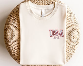 May include: A sand-colored t-shirt with a red, white, and blue "USA" graphic on the front. The graphic has a vintage, collegiate style.