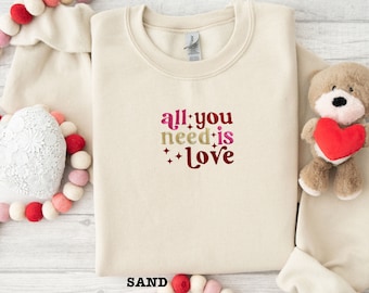 May include: A beige sweatshirt with the text "all you need is love" in a colorful, retro font. The sweatshirt is surrounded by pink and red heart-shaped decorations.