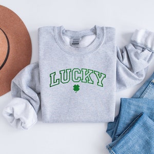 以下が含まれることがあります： 緑色の縁取りのある文字で「LUCKY」と書かれたグレーのトレーナーと緑色の四つ葉のクローバー