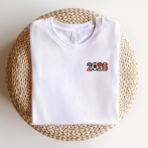 May include: White t-shirt with a small, colorful graphic of a soccer ball and the number "2026" on the front. The graphic incorporates red, white, blue, and green colors. The shirt is folded and resting on a woven, circular surface.