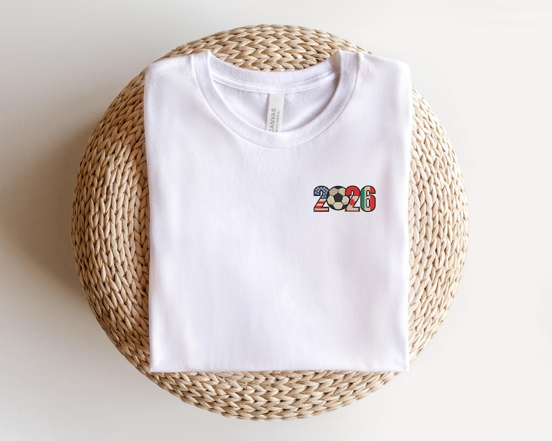 May include: White t-shirt with a small, colorful graphic of a soccer ball and the number "2026" on the front. The graphic incorporates red, white, blue, and green colors. The shirt is folded and resting on a woven, circular surface.