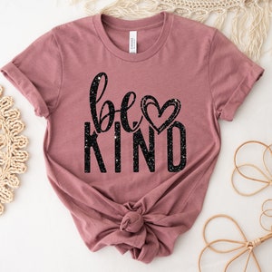 May include: A mauve t-shirt with the text "be kind" in black glitter lettering and a small heart graphic.