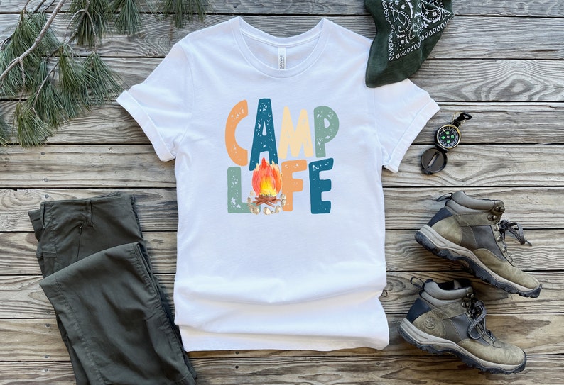 K&ouml;nnte beinhalten: Wei&szlig;es T-Shirt mit einer bunten Grafik, auf der "Camp Life" steht, mit einem Lagerfeuer in der Mitte des Wortes "Life".
