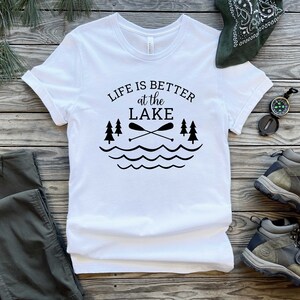 May include: White t-shirt with black text that reads "Life is better at the lake" with a graphic of two crossed paddles, three pine trees, and wavy lines representing water.