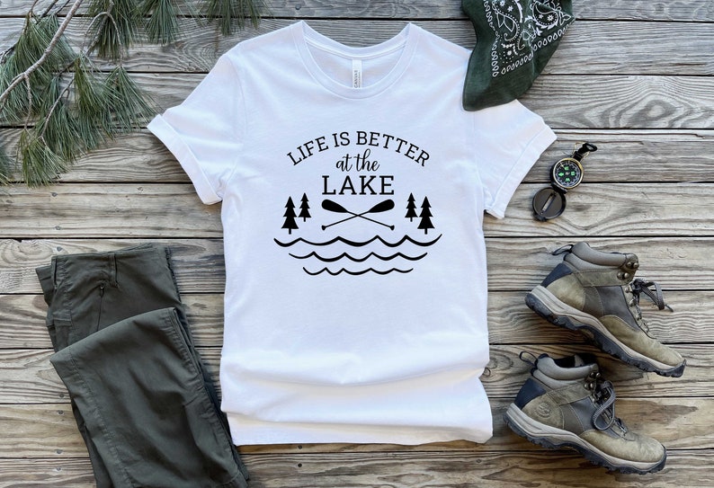 May include: White t-shirt with black text that reads "Life is better at the lake" with a graphic of two crossed paddles, three pine trees, and wavy lines representing water.