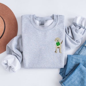 May include: A gray sweatshirt with a white crew neck and a small embroidered image of a golden retriever wearing a green shamrock. The sweatshirt is laid out on a white surface with a brown hat and blue jeans.