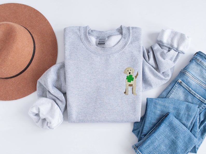 May include: A gray sweatshirt with a white crew neck and a small embroidered image of a golden retriever wearing a green shamrock. The sweatshirt is laid out on a white surface with a brown hat and blue jeans.