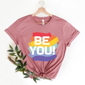 May include: A mauve t-shirt with a rainbow-colored graphic that says "BE YOU!" in white text.