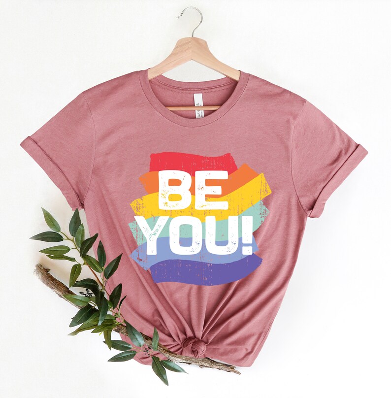 May include: A mauve t-shirt with a rainbow-colored graphic that says "BE YOU!" in white text.