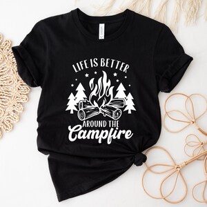 May include: Black t-shirt with a white graphic design that says "Life is better around the campfire". The graphic features a campfire with flames, logs, and trees.