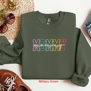 Pode incluir: Uma camisola verde militar com a palavra "MOMMY" escrita em cores do arco-&iacute;ris. A camisola &eacute; de gola redonda com mangas compridas.