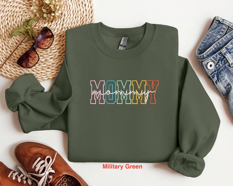 Pode incluir: Uma camisola verde militar com a palavra "MOMMY" escrita em cores do arco-&iacute;ris. A camisola &eacute; de gola redonda com mangas compridas.