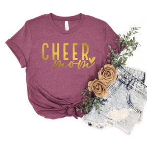 May include: A maroon t-shirt with the text "CHEER mom" in gold lettering with a small gold heart.