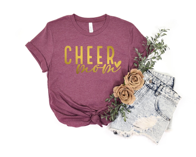 May include: A maroon t-shirt with the text "CHEER mom" in gold lettering with a small gold heart.