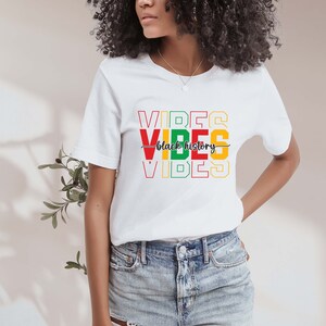 May include: White t-shirt with the words "VIBES" stacked on top of each other in red, yellow, and green. The words "black history" are written in black cursive script between the two sets of "VIBES".