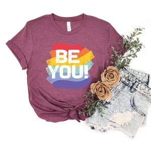 May include: A maroon t-shirt with a rainbow graphic that says "BE YOU!" in white text. The shirt is paired with denim shorts and a sprig of greenery.