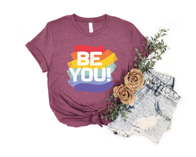May include: A maroon t-shirt with a rainbow graphic that says "BE YOU!" in white text. The shirt is paired with denim shorts and a sprig of greenery.