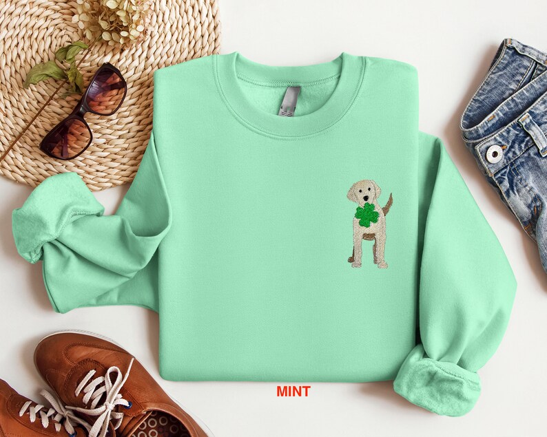 May include: A mint green sweatshirt with a small embroidered image of a golden retriever holding a four-leaf clover. The text "MINT" is printed on the bottom of the sweatshirt.