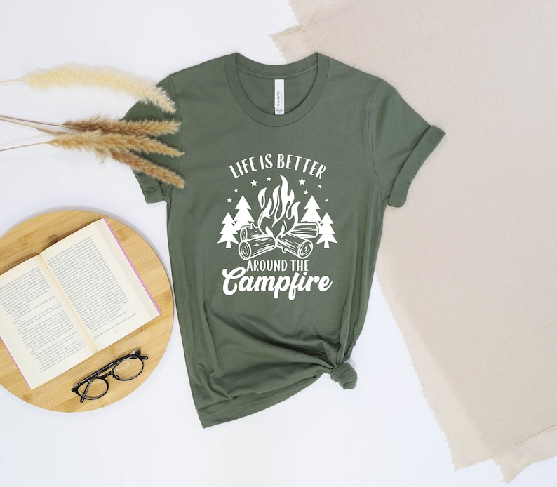 May include: A green t-shirt with a white graphic that says "Life is better around the campfire". The graphic features a campfire with flames and logs, surrounded by trees and stars.