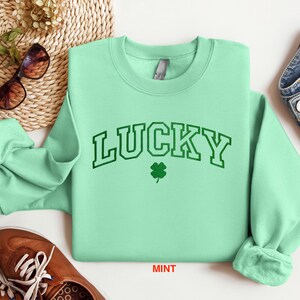 以下が含まれることがあります： ミントグリーンのトレーナーに、緑色の縁取りのある文字で「LUCKY」と書いてあり、緑色の四つ葉のクローバーが描かれています。