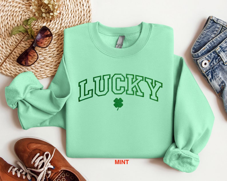 以下が含まれることがあります： ミントグリーンのトレーナーに、緑色の縁取りのある文字で「LUCKY」と書いてあり、緑色の四つ葉のクローバーが描かれています。