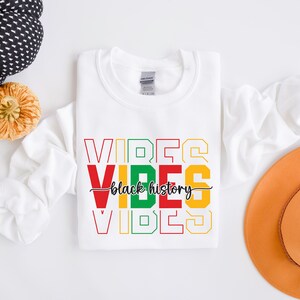 May include: White sweatshirt with the words "VIBES" in red, yellow, green, and black, stacked on top of each other. The words "black history" are written in black cursive script between the two "VIBES" words.
