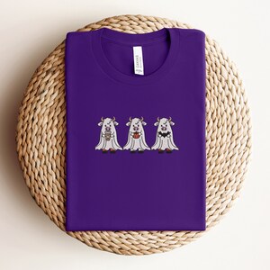 K&ouml;nnte beinhalten: Lilafarbenes T-Shirt mit drei wei&szlig;en Geisterk&uuml;hen, die Kaffeetassen und K&uuml;rbisse halten. Der Text "Canvas" befindet sich auf der R&uuml;ckseite des Shirts.