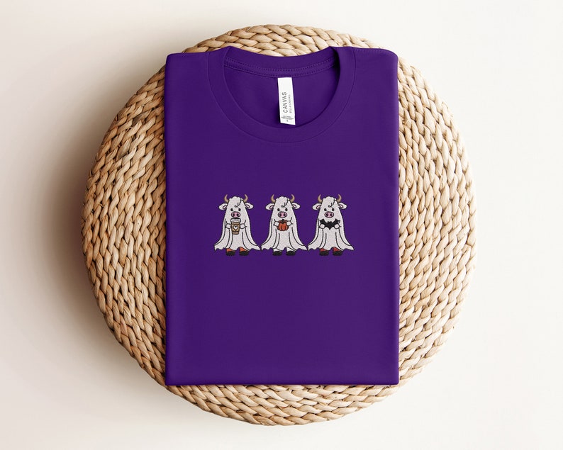 K&ouml;nnte beinhalten: Lilafarbenes T-Shirt mit drei wei&szlig;en Geisterk&uuml;hen, die Kaffeetassen und K&uuml;rbisse halten. Der Text "Canvas" befindet sich auf der R&uuml;ckseite des Shirts.