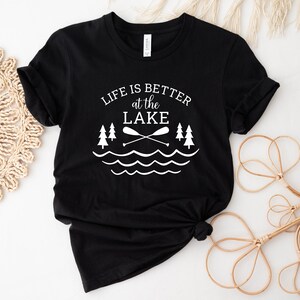 May include: Black t-shirt with white text that reads "Life is better at the lake" with a graphic of two trees, two crossed paddles, and wavy lines representing water.
