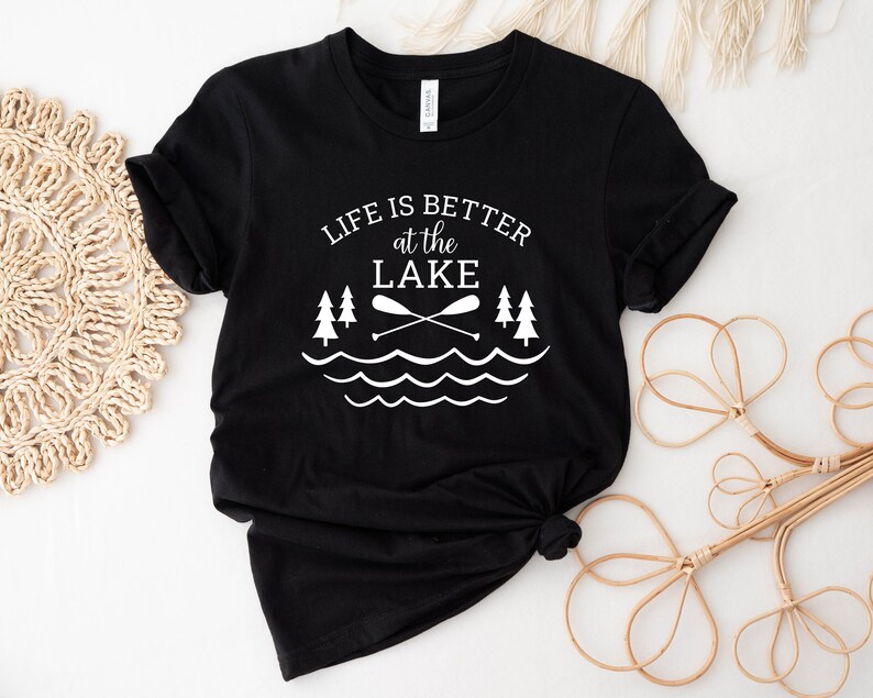May include: Black t-shirt with white text that reads "Life is better at the lake" with a graphic of two trees, two crossed paddles, and wavy lines representing water.