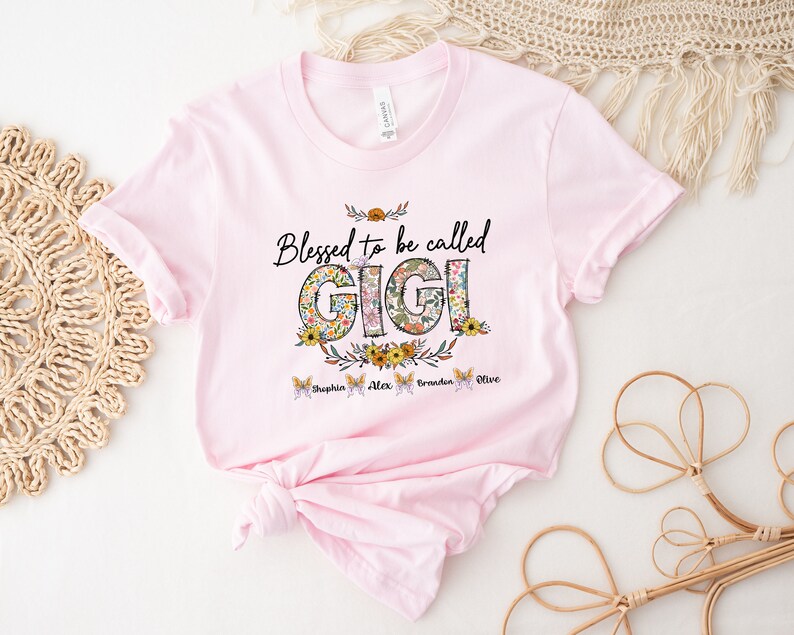 May include: Pink t-shirt with the text "Blessed to be called Gigi" in black lettering with floral accents. The names "Sophia, Alex, Braden, Olive" are written in smaller font below the text.