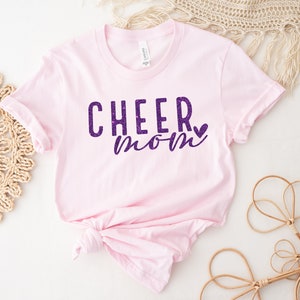 May include: Pink t-shirt with the text "CHEER mom" in purple glitter lettering with a small purple glitter heart.