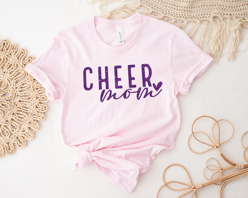 May include: Pink t-shirt with the text "CHEER mom" in purple glitter lettering with a small purple glitter heart.